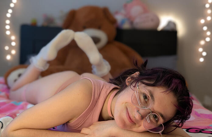 pastel is laying on their stomach on their bed with their feet up and smiling at the camera. she's wearing pink heart shaped glasses and her purple and dark brown hair is tied back into pigtails. her pink tank top matches her pink collar and her white sock